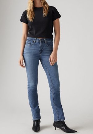 Femme portant un jean bleu droit, un top noir à manches courtes et des bottines noires à talons, debout sur un fond uni.
