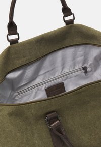 Bolso de lona en verde oliva con una correa de cuero marrón. El interior cuenta con un forro a rayas, un bolsillo con cremallera y una etiqueta de cuero.