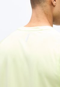 Κοντινή λήψη από την άνω πίσω πλευρά και τον λαιμό ενός ατόμου που φοράει ένα ελαφρύ κίτρινο υφασμάτινο πουκάμισο με μια μικρή αντανακλαστική λωρίδα κοντά στον γιακά.