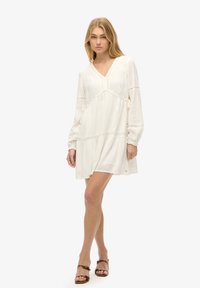 Vestido blanco, holgado, con escote en V, mangas largas y acentos de encaje. Tejido texturizado con fruncido en la cintura y una silueta relajada.