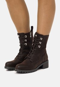 Botas de tobillo de ante marrón con cordones en la parte frontal, detalles en hardware plateado y suela de goma texturizada. Presenta una cremallera lateral para facilitar su uso.