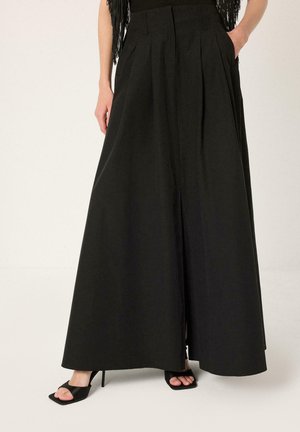 Γυναίκα που φορεί μαύρο παντελόνι ψηλόμεσο με φαρδιά μπατζάκια και πιέτες, συνδυασμένο με μαύρα πέδιλα με ψηλό τακούνι και ανοιχτά δάχτυλα, στέκεται με τα χέρια στις τσέπες.