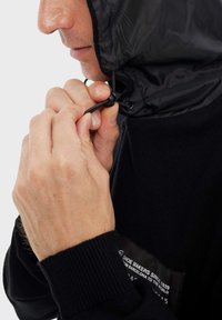 Giacca nera con cappuccio, dotata di chiusura con cordino. Polsini a costine e etichetta laterale. Primo piano di mani che regolano il cappuccio.