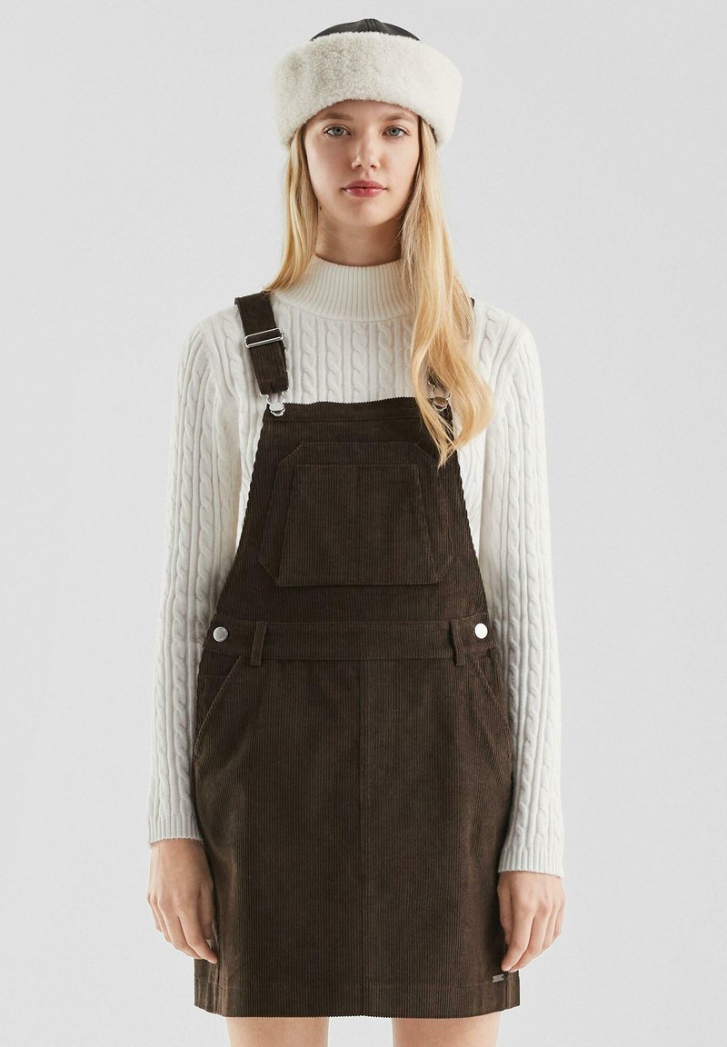 Junge Frau, die einen weißen Kabelstrickpullover und ein dunkelbraunes Cordkleid mit beigem Pelz-Haarband trägt, steht vor einem schlichten Hintergrund.