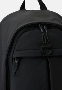 Gros plan d'un sac à dos noir montrant la poche zippée avant avec deux tirettes et un petit détail de boucle noire au-dessus de la poche.