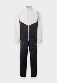 Σετ φόρμας Nike με ζακέτα με φερμουάρ, ανοιχτό γκρι στην πάνω πλευρά και μαύρο κάτω μέρος, συνοδευόμενο από μαύρο παντελόνι. Πράσινες λεπτομέρειες με λογότυπο.