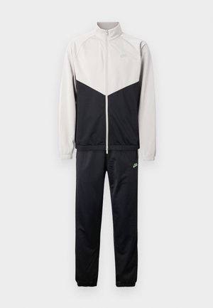 Dres Nike z kurtką na zamek błyskawiczny, która ma górną część w jasnoszarym kolorze i dolną w czarnym, w zestawie z czarnymi spodniami. Zielone akcenty z logo.