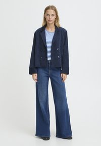 Veste croisée en velours côtelé bleu marine portée sur une chemise rayée bleu et blanc, associée à un jean large en denim foncé.