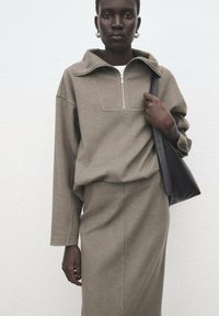 Szara, oversize bluza z kołnierzem na suwak do ćwiartki, zestawiona z dopasowaną spódnicą. Modelka trzyma czarną torebkę typu tote ze skóry. Gładka faktura tkaniny.