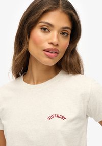 Beżowa bawełniana koszulka z krótkim rękawem, z wyhaftowanym na lewej piersi zakrzywionym czerwonym logo "SUPERDRY". Gładka faktura.