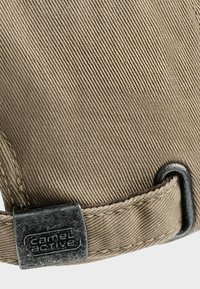 Beige corduroy stof med en subtil tekstur, prydet med et sort metal logo mærke og en kontrasterende syet kant.