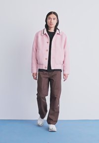Jovem com cabelo longo e escuro, vestindo uma jaqueta rosa, camisa preta, calças castanhas e ténis brancos, a caminhar sobre um chão azul contra uma parede branca.