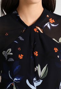 Blouse marine avec un détail noué au niveau du col, motifs floraux en orange, vert et bleu, réalisée en tissu léger et transparent.