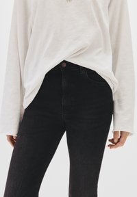 Ein langärmliges weißes Oberteil fällt über dunkle hochgeschnittene Jeans. Die Jeans haben eine glatte Textur und ein einzelnes, rundes Knopfdetail.