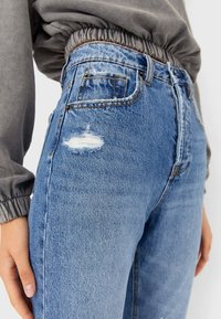 Jeans de mezclilla azul de talle alto con un acabado descolorido y ligero desgaste. Cuenta con cinco bolsillos y costuras en contraste. Textura suave.
