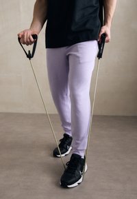 Purple sweatpants and black athletic shoes, holding resistance bands with black handles, standing on a neutral floor against a beige wall.