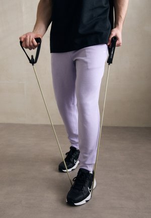 Pantalons de survêtement violets et chaussures de sport noires, tenant des bandes de résistance avec des poignées noires, debout sur un sol neutre contre un mur beige.