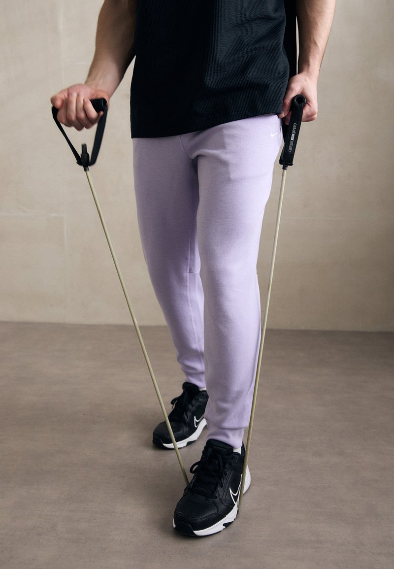 Purple sweatpants and black athletic shoes, holding resistance bands with black handles, standing on a neutral floor against a beige wall.