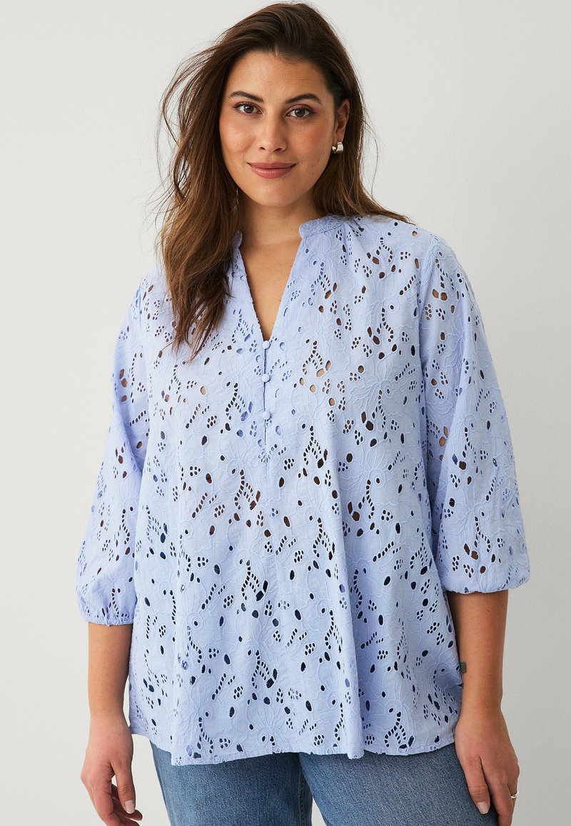 Hellblaue Bluse aus bestickter Baumwolle mit floralen Ausschnitten, V-Ausschnitt und Dreiviertelärmeln, kombiniert mit Denim.