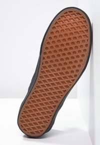 Semelle d'une chaussure noire avec une semelle en caoutchouc brun à motif gaufré, appuyée contre un mur et un sol blancs.