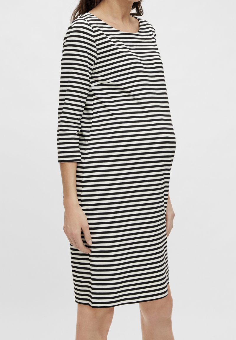 Femme enceinte portant une robe noire et blanche à rayures horizontales, longueur genou, manches trois-quarts, debout de profil.