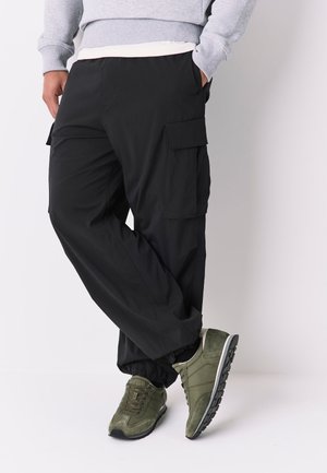 Persona che indossa pantaloni cargo neri con tasche laterali, felpa grigia e scarpe da ginnastica verdi, in piedi con le gambe incrociate.