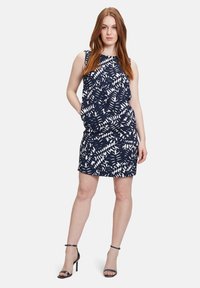 Ärmelloses Kleid in Marineblau mit einem weißen Blättermuster, das eine lockere Passform und ein schichtartiges Design aufweist. Kombiniert mit Riemchen-High Heels.