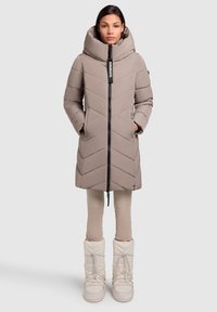 Beige pufferjakke med chevronmønster, stor hætte og en lynlås foran. Sammen med lyse beige leggings og hvide, isolerede støvler.