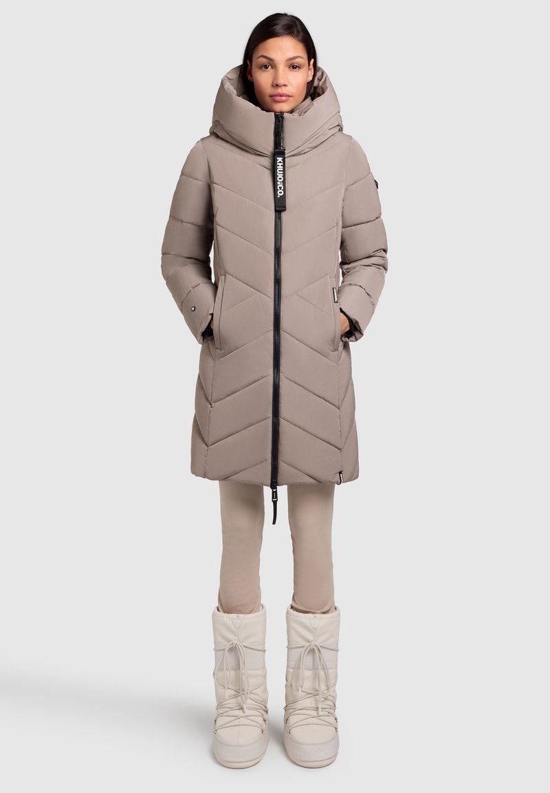 Beige pufferjakke med chevronmønster, stor hætte og en lynlås foran. Sammen med lyse beige leggings og hvide, isolerede støvler.