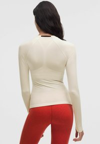 Camiseta de manga larga y cuello redondo en tejido crema, con un diseño ajustado, mangas raglán y un pequeño logo en la parte posterior cerca del escote.