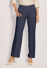 Vrouw in marineblauwe rechtliggende broek, beige puntneus hoge hakken, crèmekleurig getextureerd topje en gouden armband om de linkerpols.