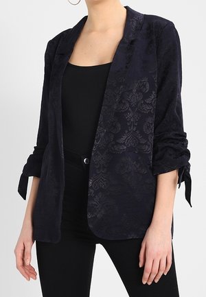 Kobieta ubrana w ciemną, wzorzystą marynarkę z marszczonymi rękawami związanymi przy mankietach, w połączeniu z czarnym, bezrękawnym topem i czarnymi spodniami.