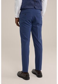 Mężczyzna w dopasowanych niebieskich spodniach, ciemnych brązowych butach eleganckich, białej koszuli i granatowej kamizelce, stojący na jednolitym tle.