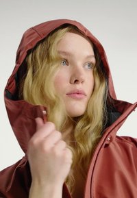 Junge Frau mit langen blonden Haaren, die eine rostrote Kapuzenjacke trägt, hält den Reißverschluss und schaut leicht zur Seite.