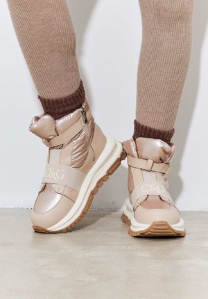 Beige gewatteerde enkelboots met een glanzende afwerking, elastische band met logo, zijrits en witte zool met een gestructureerde bruine buitenzool.