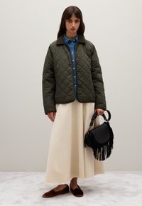 Junge Frau in einer gesteppten olivgrünen Jacke über einem blauen Shirt, einer langen cremefarbenen Rock, braunen Ballerinas und hält eine schwarze Fransenhandtasche.