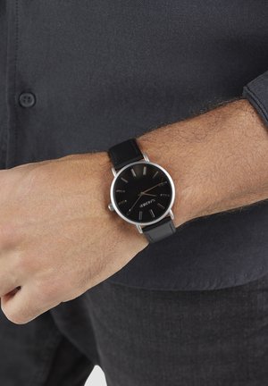 Schwarze Armbanduhr mit rundem Zifferblatt, silbernen Elementen und schwarzem Lederarmband. Die Uhr verfügt über ein schlichtes schwarzes Zifferblatt mit minimalistischen silbernen Stundenmarkierungen.