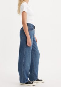 Žena v bílém krátkém tričku s krátkým rukávem, v volných modrých džínách s vysokým pasem a černých nízkých teniskách, stojící na bílém pozadí, boční pohled.