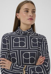 Top de cuello alto azul marino con un patrón geométrico blanco que incluye cuadrados y círculos. Tejido suave con mangas largas y diseño ajustado.