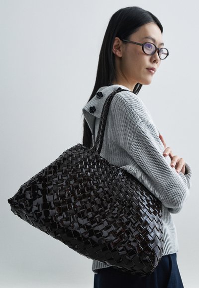 Geslagen zwarte tas met een glanzende afwerking, voorzien van een ruim ontwerp en twee stevige handvatten, getoond naast een lichtgrijze trui.