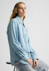 Camicia in denim blu chiaro con maniche lunghe, bottoni frontali e taschino sul petto. Presenta dettagli strappati e una vestibilità rilassata.