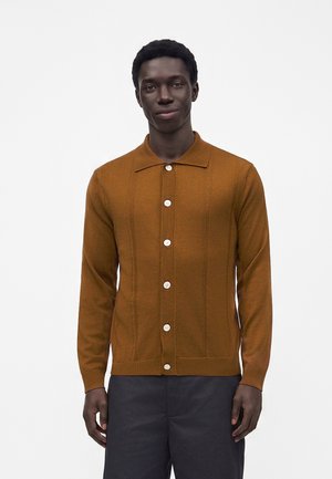 Homme portant un cardigan boutonné couleur rouille avec des boutons blancs et un pantalon foncé, debout devant un fond clair uni.