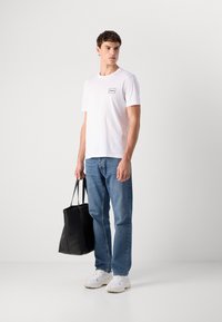 Hvit bomulls t-skjorte med svart logo, kombinert med blå denimjeans og hvite joggesko; holder en svart totebag med doble håndtak.