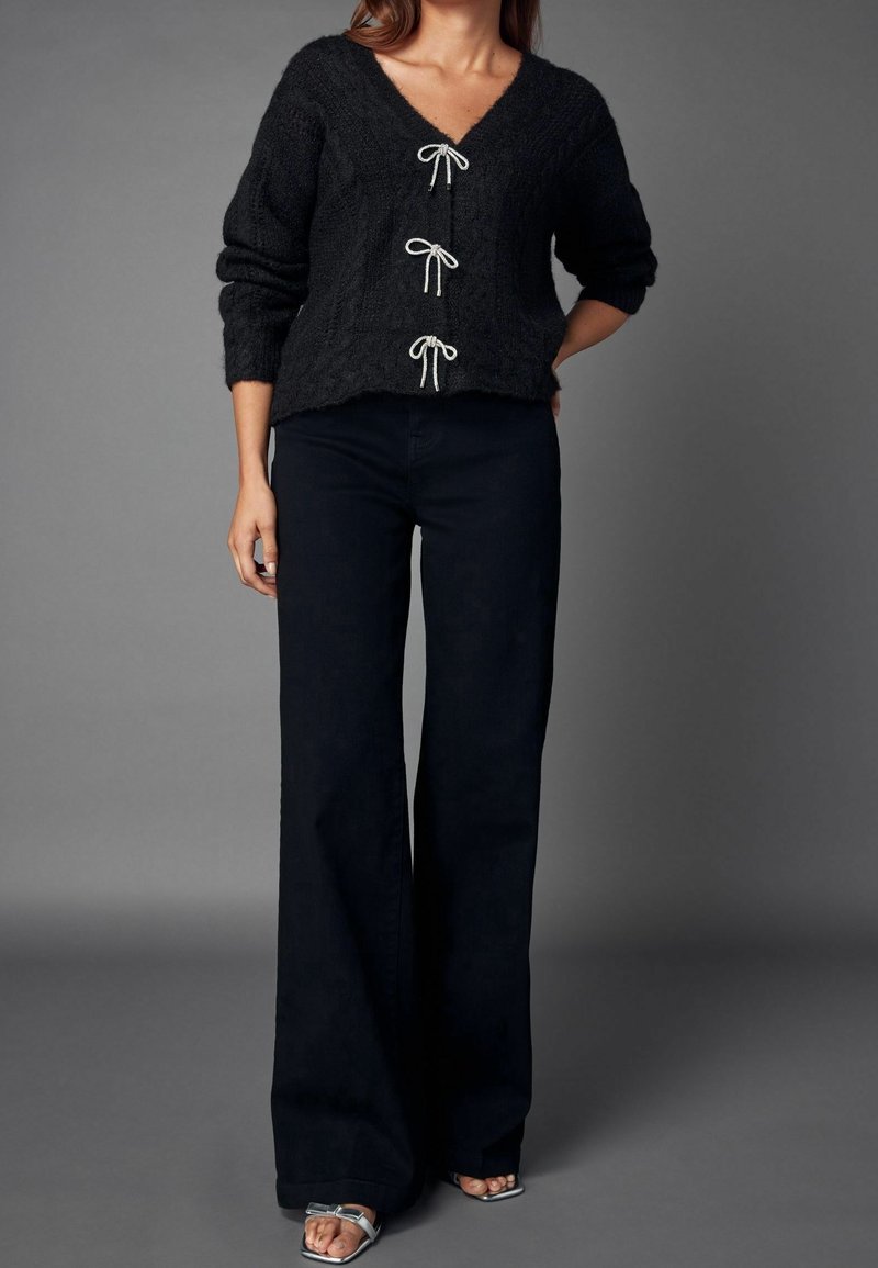 Femme portant un cardigan noir en maille avec trois nœuds blancs, un pantalon noir à jambes larges et des sandales argentées à bout ouvert sur fond gris.