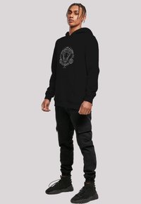 Juodas hoodie su centrinėmis grafikos detalėmis, vaizduojančiomis liūto galvą, papildytas apskritimais. Pagamintas iš švelnios medžiagos, turi kengūros stiliaus kišenę.
