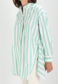 Žena nosící dlouhou bílou košili na knoflíky s vertikálními zelenými pruhy a dlouhými rukávy, v kombinaci s světlými kalhotami.