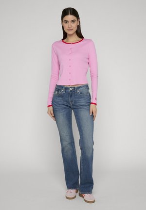 Kobieta ubrana w dopasowany różowy kardigan z czerwonym wykończeniem, niebieskie dżinsy i jasnoróżowe trampki, stojąca na tle jednolitego szarego tła.