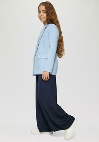 Jasnoniebieska marynarka o strukturalnym kroju, zestawiona z szerokimi granatowymi spodniami. Oba elementy mają gładką teksturę; strój dopełniają białe sneakersy.
