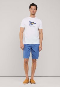 Camiseta blanca de manga corta con un gráfico náutico en azul y amarillo, combinada con pantalones cortos azules y zapatos de hombre marrones sin cordones. Diseño sencillo.