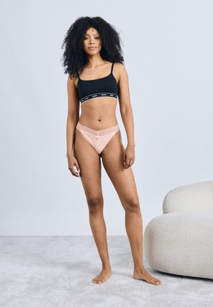 Černá braletka s páskem s logem a růžová tanga lemovaná krajkou s mašličkou. Modelka stojí na světle šedém koberci vedle měkkého, zaobleného křesla.